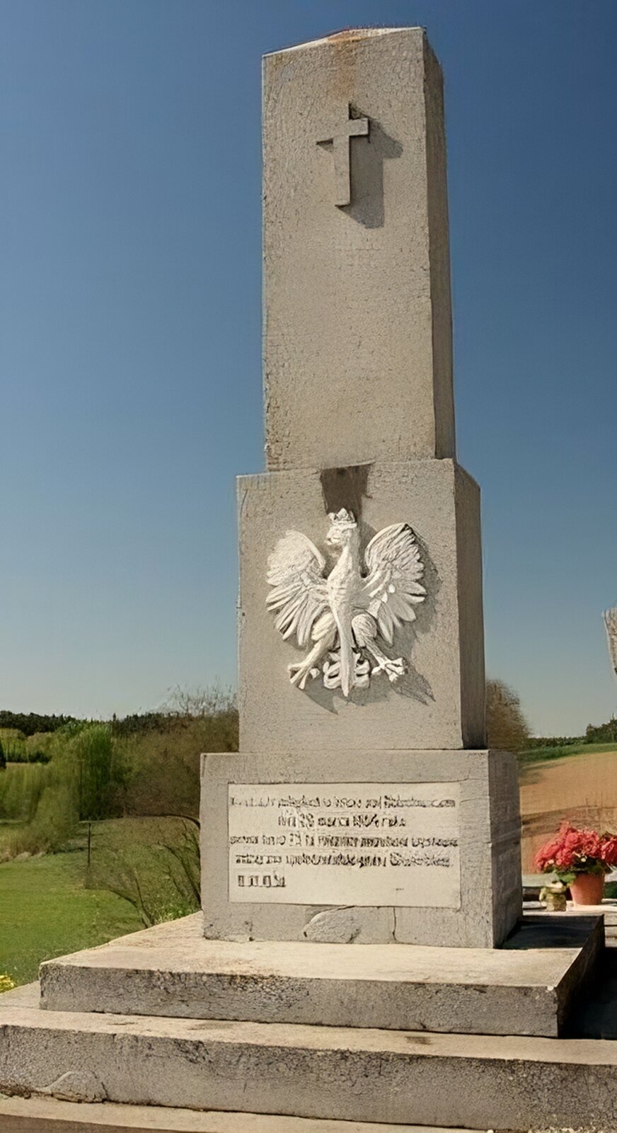 Zdjęcie przedstawia kamienny obelisk. W górnej części pomnika został wyryty krzyż. Niżej widoczny jest biały orzeł. Poniżej znajduje się pamiątkowa tablica z czarnymi napisami, które nie są czytelne. Do obelisku prowadzą dwa kamienne schody. W tle widoczna jest zielona trawa oraz drzewa i krzewy. Niebo jest błękitne, bezchmurne. Zdjęcie zostało wykonane w świetle dziennym.
