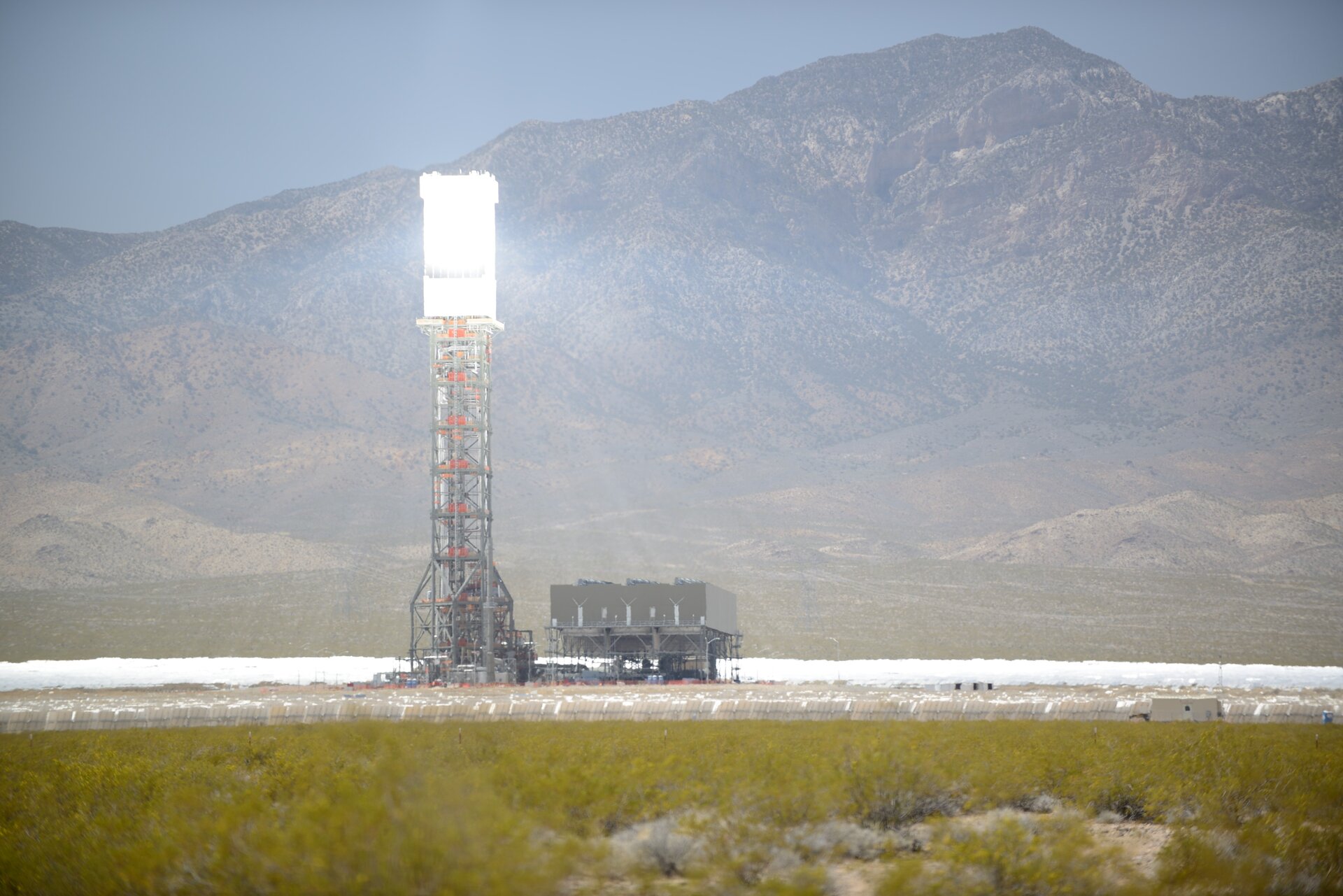 Zdjęcie przedstawia elektrownię słoneczną Ivanpah w USA. Elektrownia znajduje się w polu, w tle widać góry. Na terenie rozmieszczone są panele skierowane pod pewnym kątem względem płaszczyzny poziomej. Nieco po lewej stronie znajduje się mała platforma z zabudowaniami dla pracowników elektrowni. Obok niej stoi wieża skonstruowana z kratownicy ułożonej w prostopadłościenny kształt. Na jej szczycie zamontowany jest element odbijający światło słoneczne.