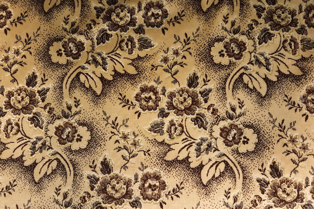 Zdjęcie ukazuje próbkę gobelinu, wzorzystego materiału. Na jasnobrązowym tle widoczny jest wzór kwiatowy w znacznie ciemniejszym odcieniu brązu.