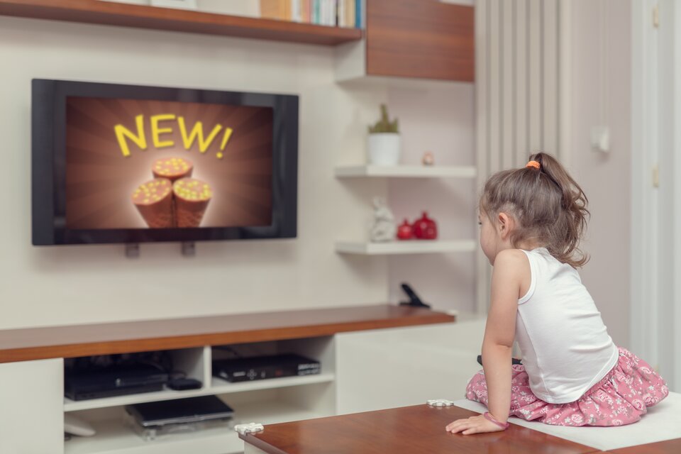 Ilustracja przedstawia dziecko zdjęcie dziewczynki siedzącej w pokoju przed telewizorem, która ogląda reklamę lodów.