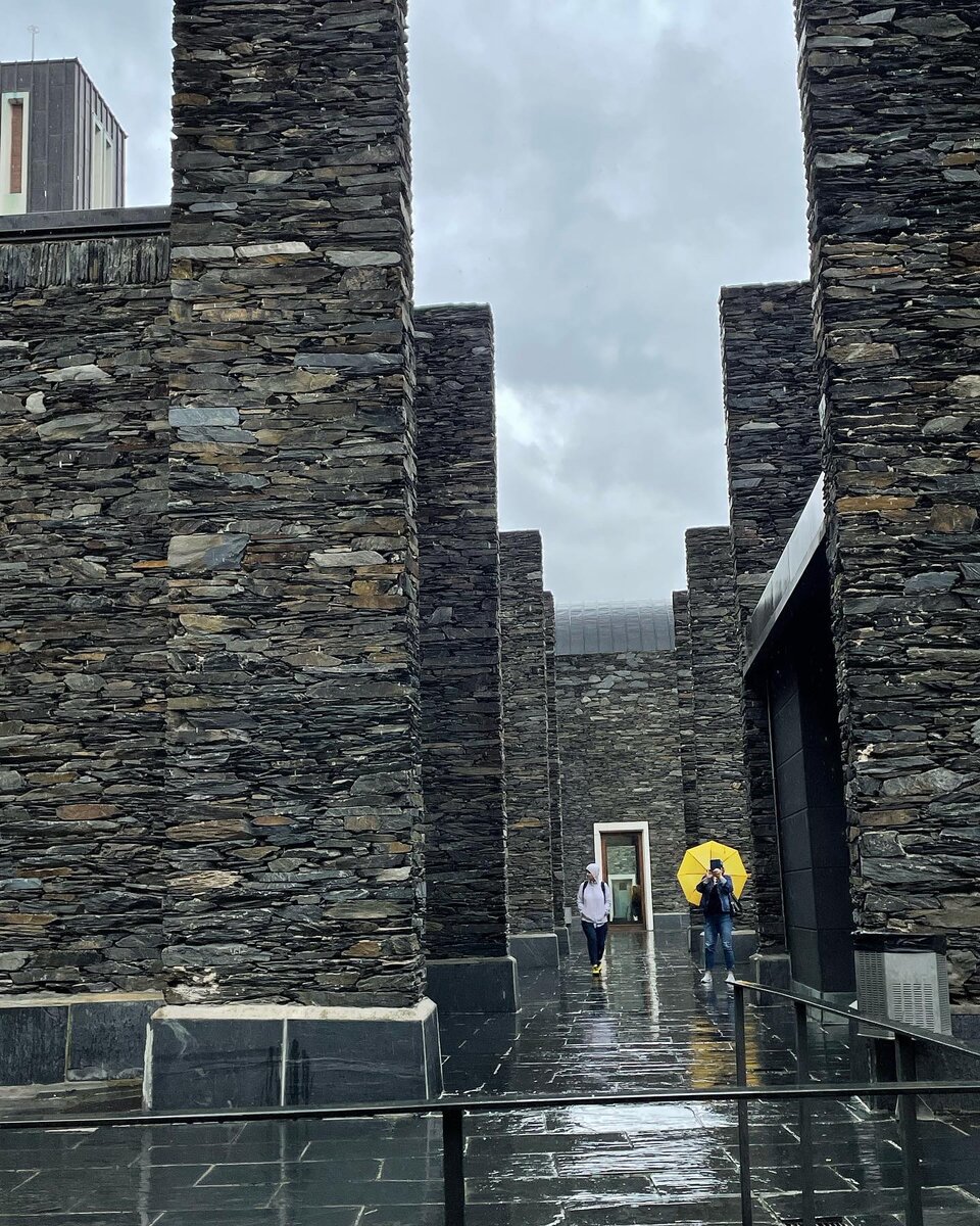 Ilustracja przedstawia fotografię wykonaną w deszczowy dzień. Fotografia przedstawia kadr z pionowymi, wysokimi kamiennymi ścianami. Pomiędzy nimi jest droga, na której znajdują się dwie postaci z parasolem. 