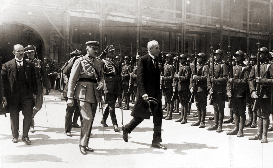 Zdjęcie przedstawia idących mężczyzn naprzeciwko stojących na baczność żołnierzy z bronią. Na czele idą dwaj mężczyźni: mężczyzna w mundurze wojskowym oraz mężczyzna w garniturze.