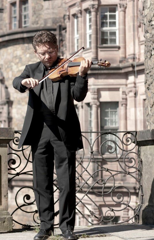 Ilustracja przedstawia mężczyznę grającego na skrzypcach na tle zabytkowego budynku.