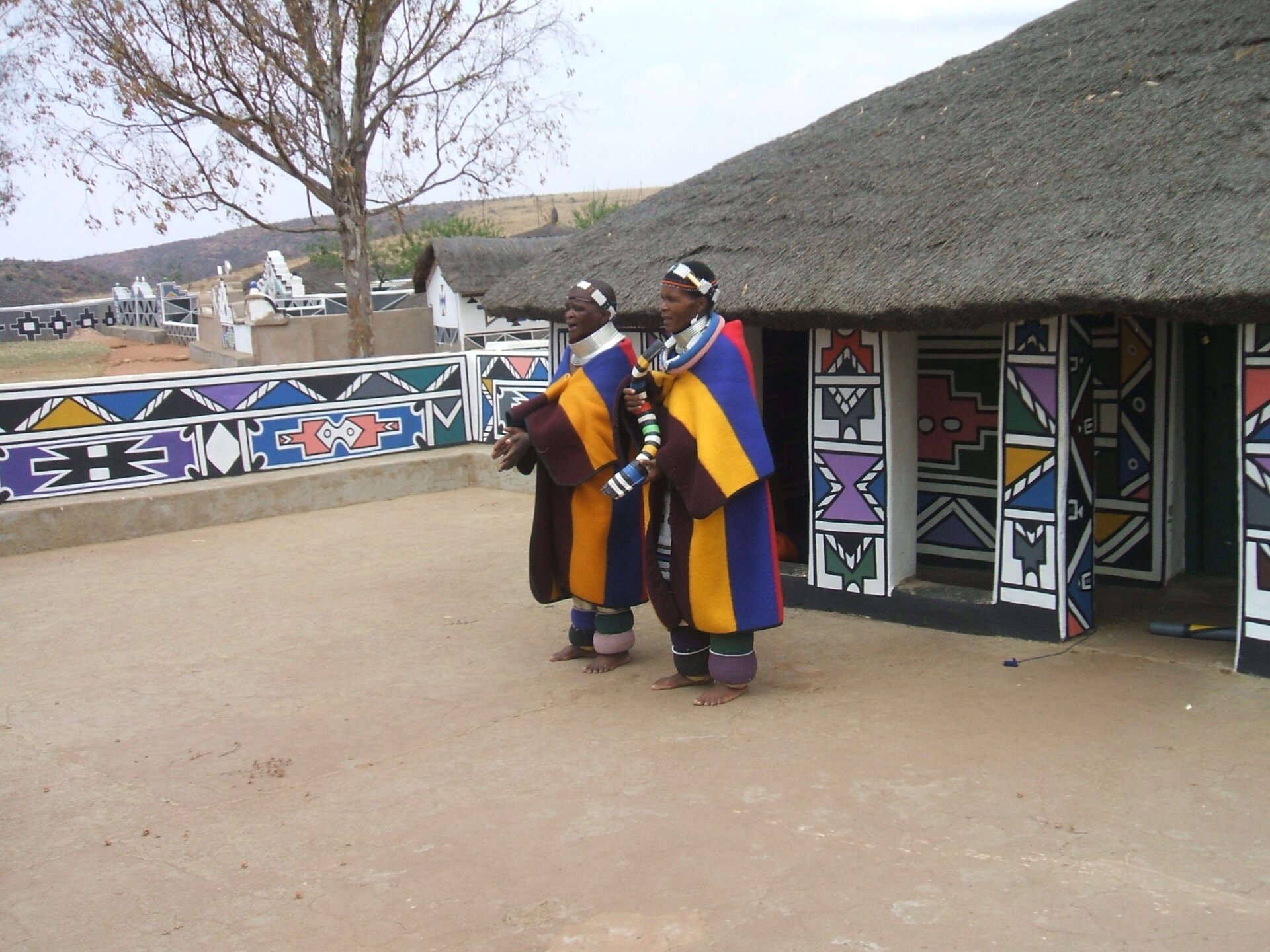 Na zdjęciu znajdują się ciemnoskórzy mieszkańcy Republiki Południowej Afryki w ludowych strojach. Ubrani są w długie suknie przypominające krojem kimona, z długimi szerokimi rękawami w kolorowe szerokie pasy - brązowy, żółty, niebieski, czerwony. Na głowach mają wąskie białe opaski. W tle znajduje się chata kryta strzechą. Jej ściany pomalowane w jaskrawe, kolorowe, geometryczne wzory. Podobne wzory widoczne są na ogrodzeniu po lewej stronie zdjęcia. Dalej widoczne jest duże drzewo i pagórkowaty teren. Niebo jest jasnoszare.