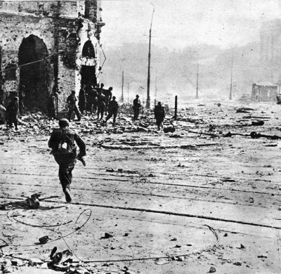 Czarno‑białe zdjęcie przedstawia żołnierza w ciemnym mundurze i hełmie, który biegnie w stronę ruin budynku. Trzyma on w ręku karabin. Przy ruinach stoją inni żołnierze. Na drodze, przez którą biegnie znajdują się tory tramwajowe oraz liczne odłamki. W oddali za kłębami dymu znajdują się budynki. 