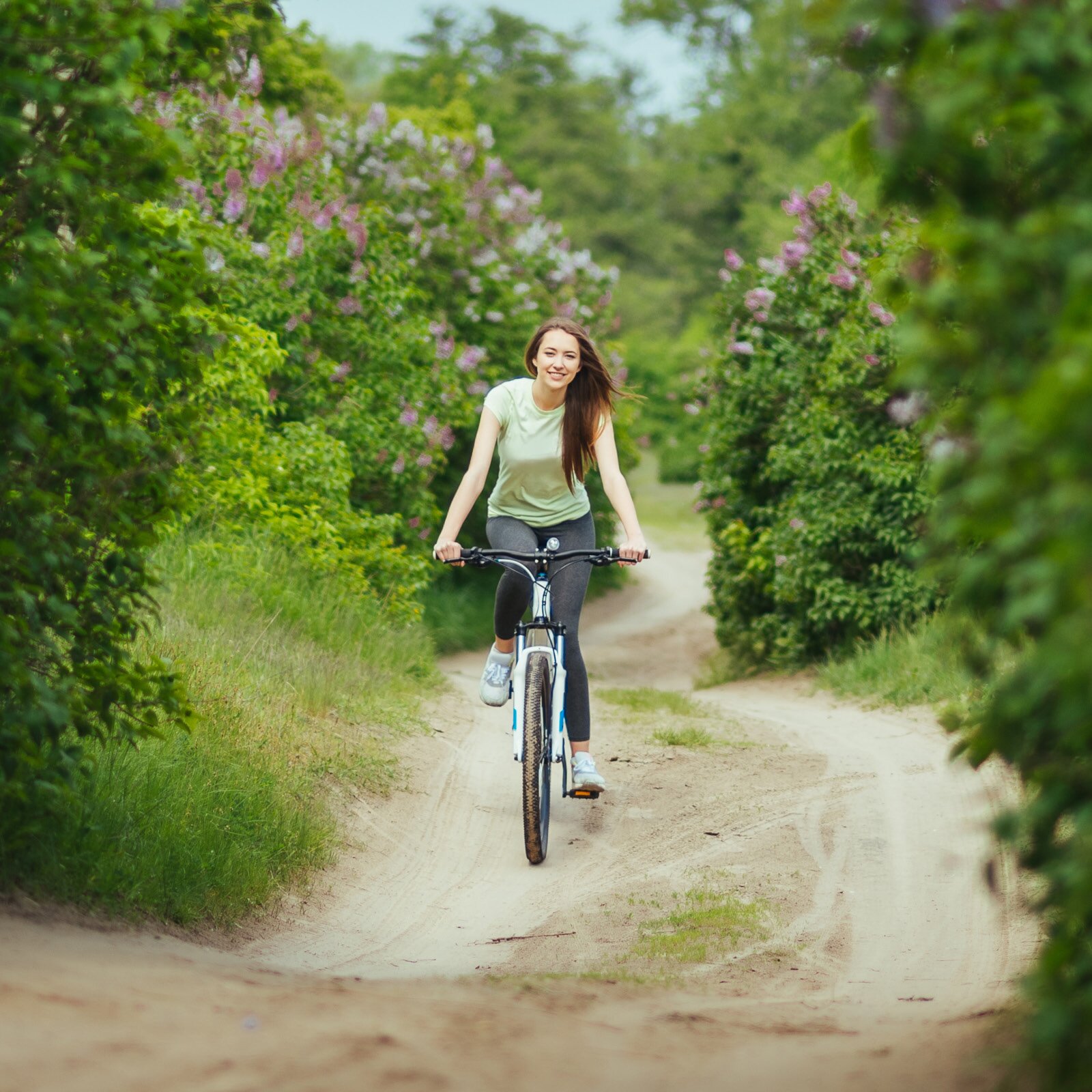 Zdjęcie przedstawia młodą kobietę jadącą na rowerze po ścieżce między krzakami bzu.