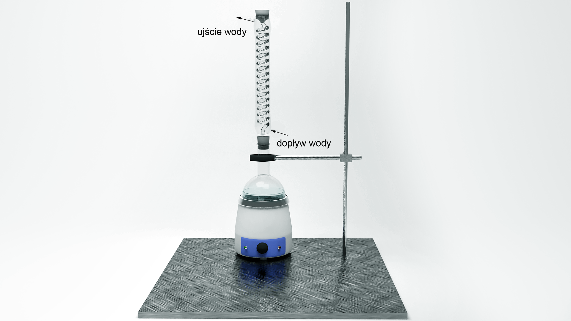 Ilustracja przedstawia następujący zestaw laboratoryjny. W łapie umieszczono kolbę okrągłodenną znajdującą się w płaszczu grzejnym, którą umocowano do statywu. W szyjkę kolby włożono chłodnicę zwrotną, usytuowaną pionowo względem blatu laboratoryjnego. Zaznaczono również dopływ wody do chłodnicy u jej dołu, a ujście u góry.