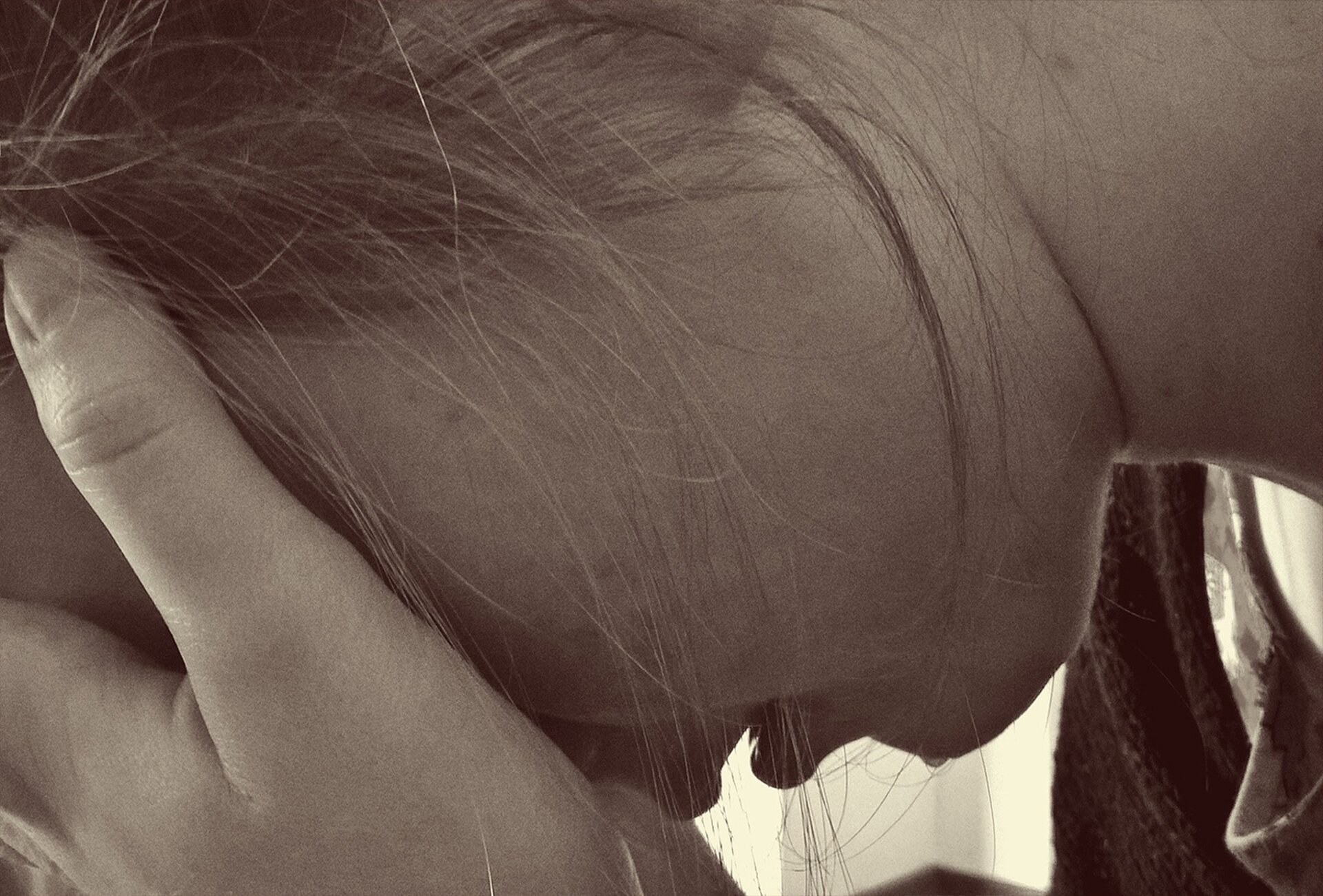 Czarno-białe zdjęcie przedstawia zbliżenie profilu długowłosej dziewczyny, która ukrywa twarz w dłoniach. Po jej brodzie spływa łza.