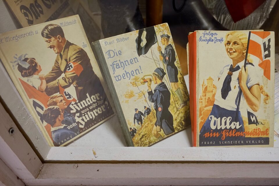 Zdjęcie przedstawia niemieckie książki na wystawie księgarni. Na okładce pierwszej książki od lewej widoczny jest Hitler z profilu, który bierze na ręce małą, niemiecką dziewczynkę z półdługimi włosami i białą kokardą. Na jego rękawie znajduje się czerwona opaska z czarną swastyką na białym tle, podobnie jak na flagach, które znajdują się obok. W środku widoczna jest okładka książki z dwoma chłopcami w mudnurach  Hitlerjugend  - jeden z nich gra na trąbce, drugi trzyma w ręce flagę. Ostatnia okładka książki przedstawia uśmiechnietą, młodą dziewczynę o spiętych z tyłu blond włosach, w białej bluzce, niebieskiej spódnicy i niebieskiej chustce pod szyją. W ręce trzyma biało – czerwoną flagę ze swastyką. 
