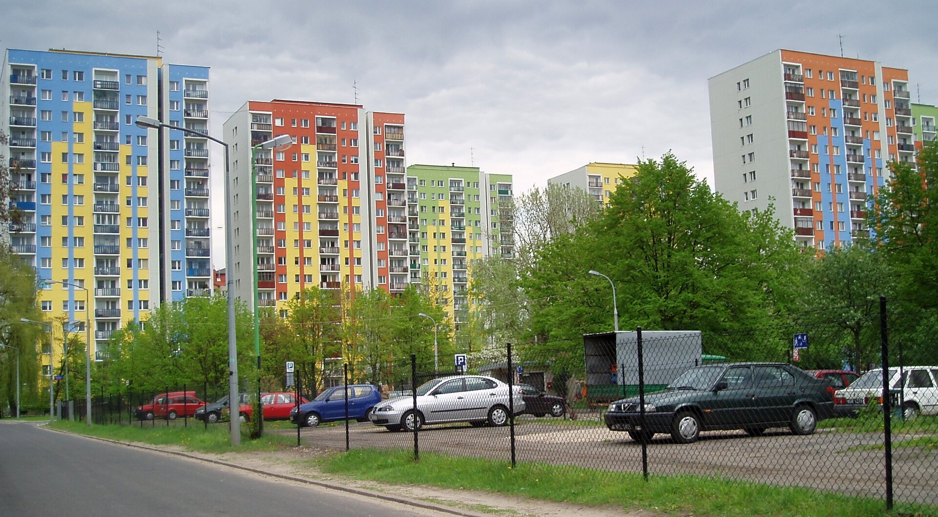 Zdjęcie przedstawia blokowisko. Każdy z widocznych wysokich budynków pomalowany jest na jaskrawe kolory. Pierwszy z nich jest żółto‑niebieski, drugi żółto‑czerwony, trzeci żółto‑zielony, czwarty niebiesko‑czerwony. Przed blokami rosną drzewa i znajduje się niewielki parking. 