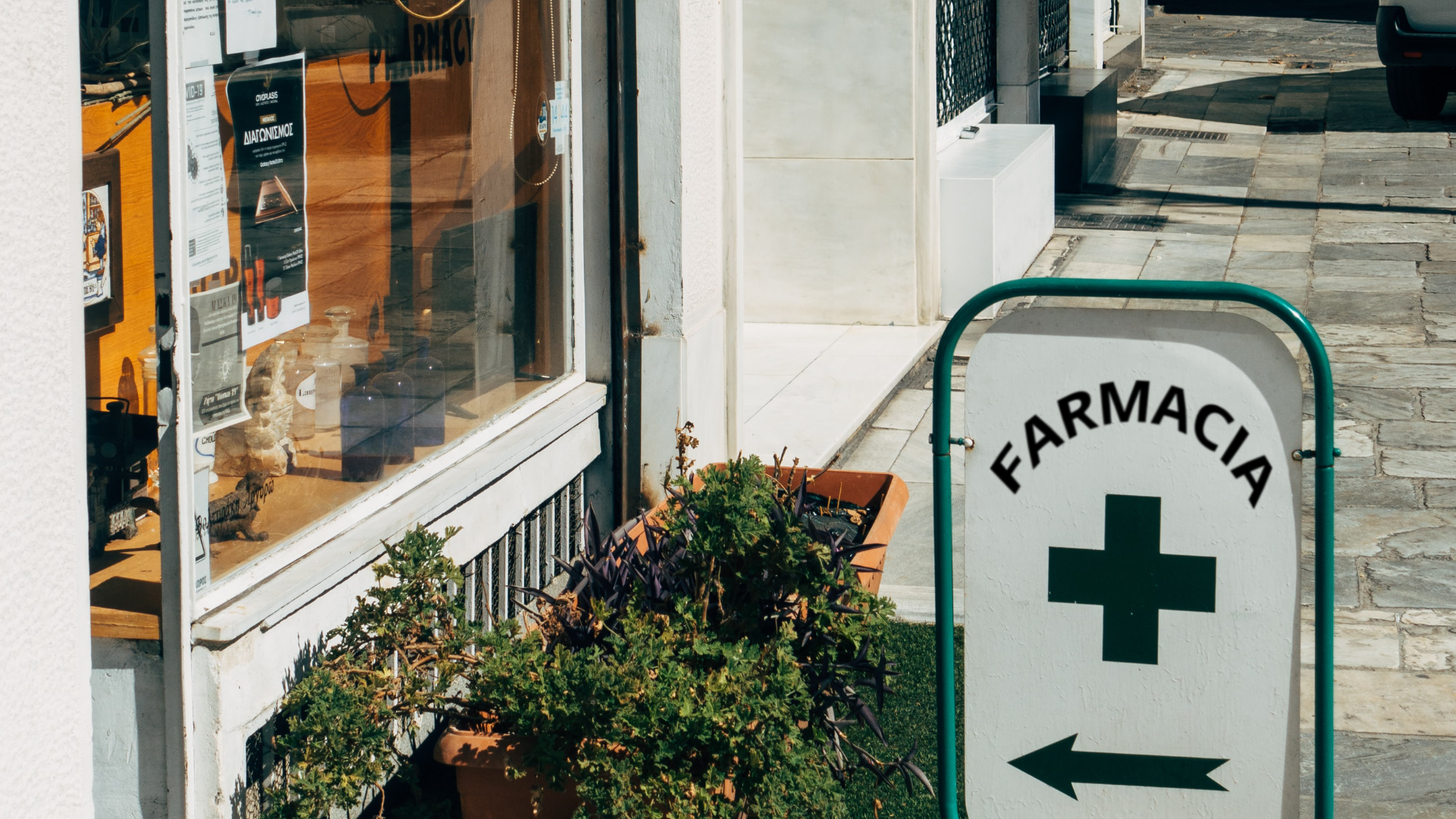 Zdjęcie przedstawia fragment fasady budynku. Przed budynkiem stoi baner z napisem "Farmacia". Na banerze znajduje się też krzyż i strzałka wskazująca wejście do apteki.