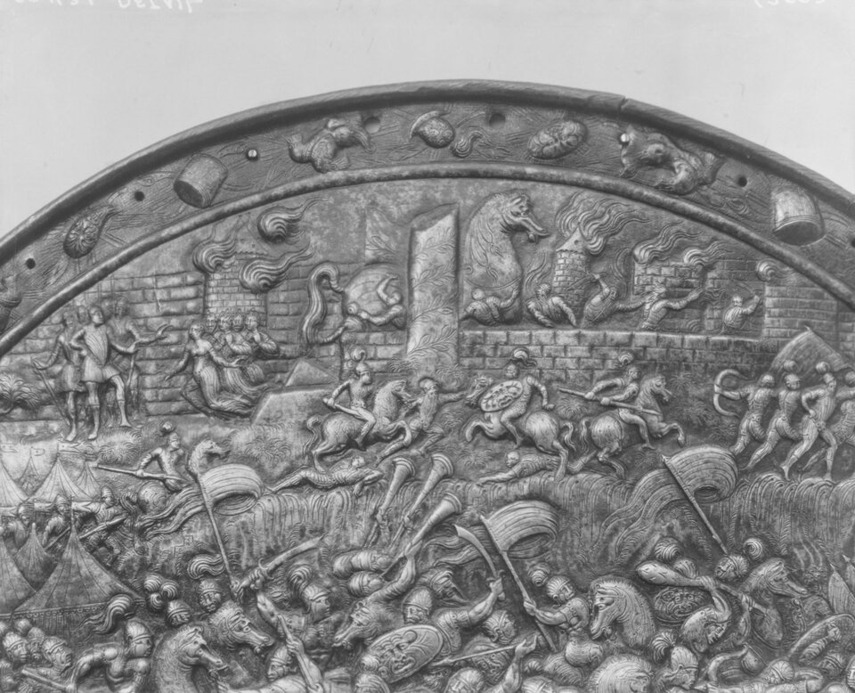 Ilustracja przestawia górny fragment tarczy wykonanej z metalu, na której znajdują się rzeźbienia. Przedstawiają one oblężenie Troi autorstwa nieznanego. Tarcza ukazuje bitwę odbywającą się pod murami miasta. Na środku kłębią się walczący wojownicy na koniach. Trzymają w ręku chorągwie, tarcze i miecze. W górnej części tarczy przestawiono mury Troi, pod które wojownicy wprowadzają konia trojańskiego.