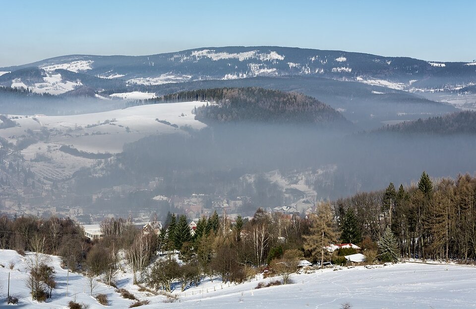 Zdjęcie obrazuje smog zalegający nad Nową Rudą położoną w dolinie rzeki Włodzicy w Sudetach. Na pierwszym planie znajduje się stok pokryty śniegiem oraz drzewami, w dolinie liczne zabudowania i drzewa przykryte szarym, nieprzejrzystym obłokiem. Ponad tym obłokiem, na dalszym planie znajduje się wzgórze porośnięte drzewami i pokryte śniegiem. 