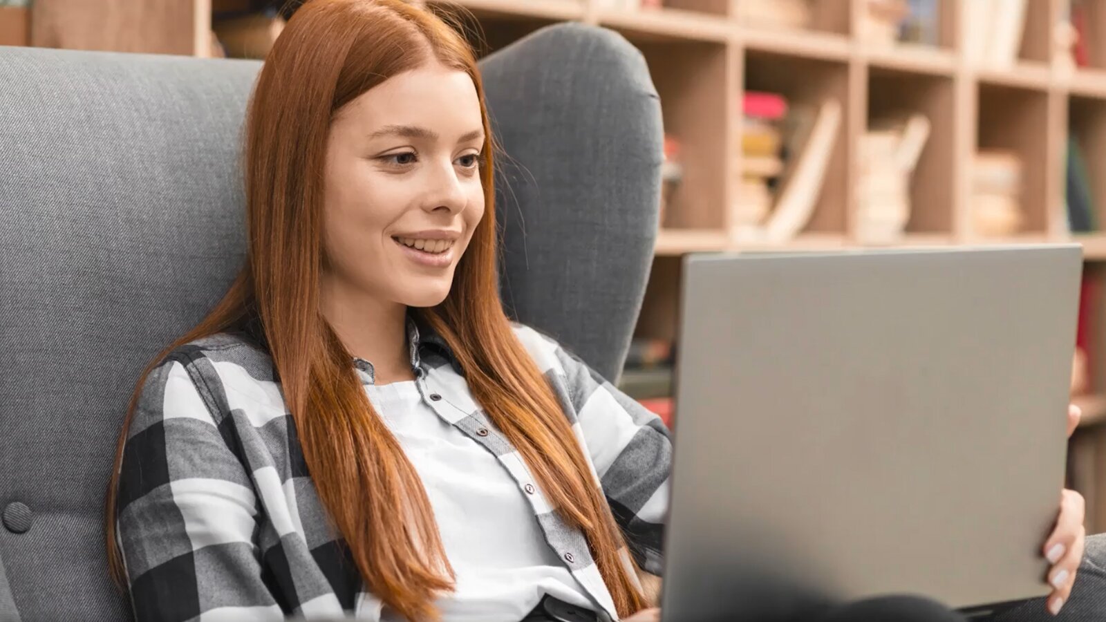 Na zdjęciu widoczna jest młoda dziewczyna wpatrująca się z uśmiechem w ekran laptopa.