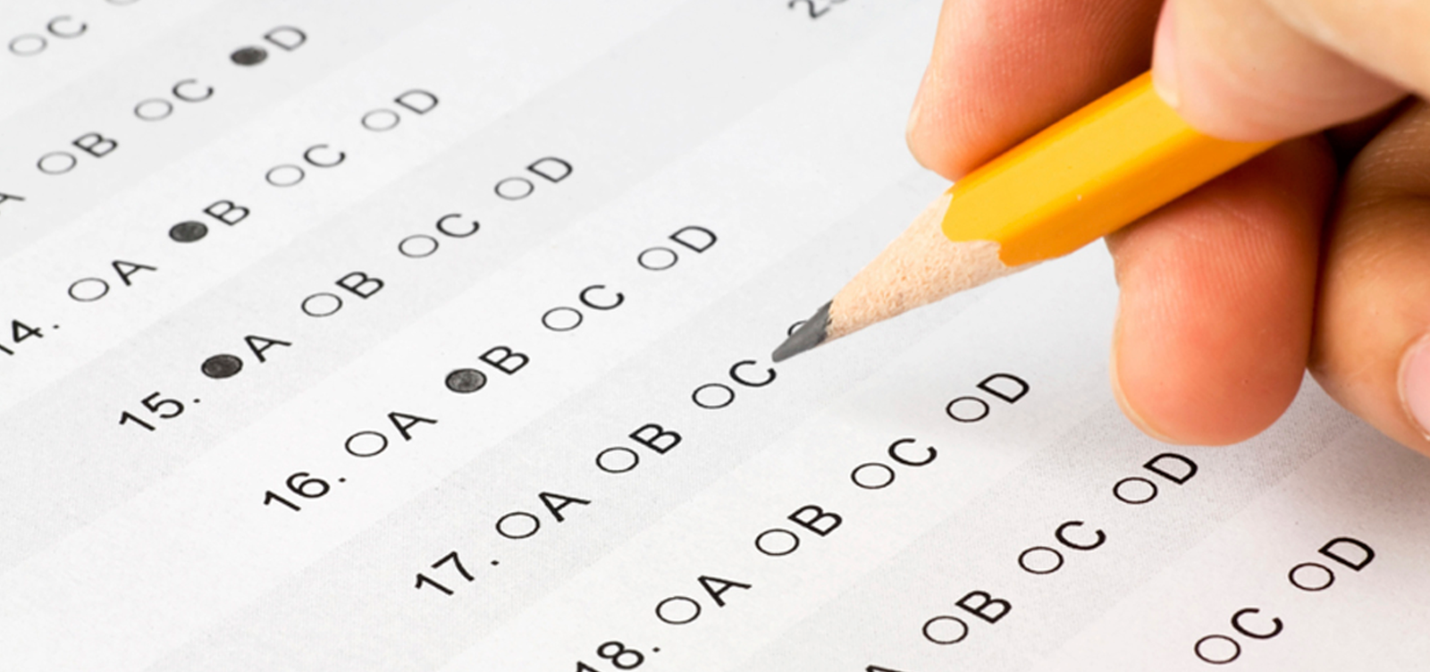 Kliknij, aby powiększyć Fotografia przedstawia białą kartkę z wydrukowanym testem wyboru odpowiedzi A, B, C, D kilku numerowanych zadań. Nad nią zatemperowany żółty ołówek trzymany w palcach prawej dłoni. Część kółek przy literach odpowiedzi kolejnych pozycji testu została zaznaczona ołówkiem.