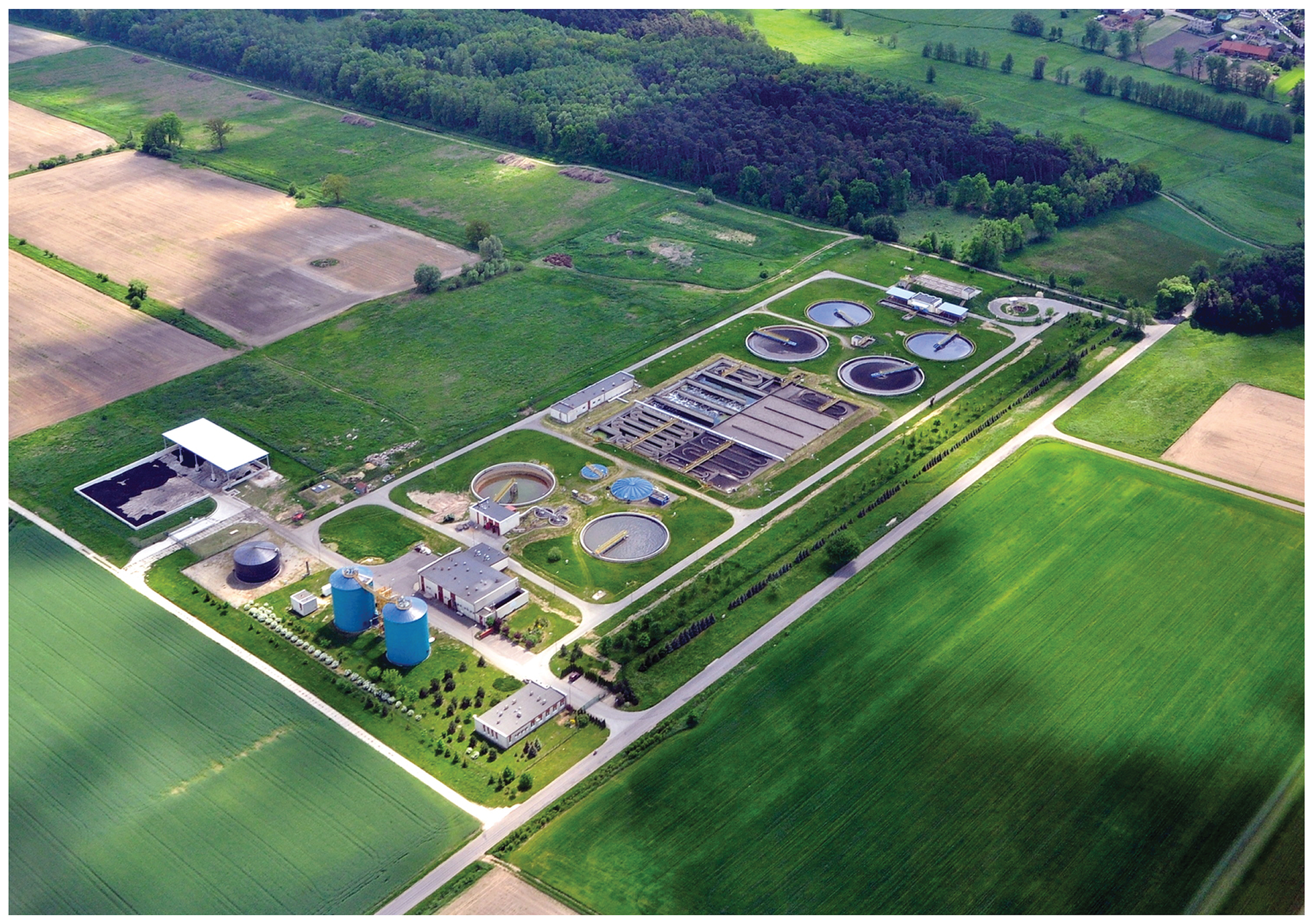 Fotografia przedstawia budowlę techniczną, widzianą z góry. Znajdują się tam cztery okrągłe zbiorniki, dwa silosy i wiele budynków. Jest to biologiczna oczyszczalnia ścieków. Tu bakterie rozkładają organiczne składniki ścieków.