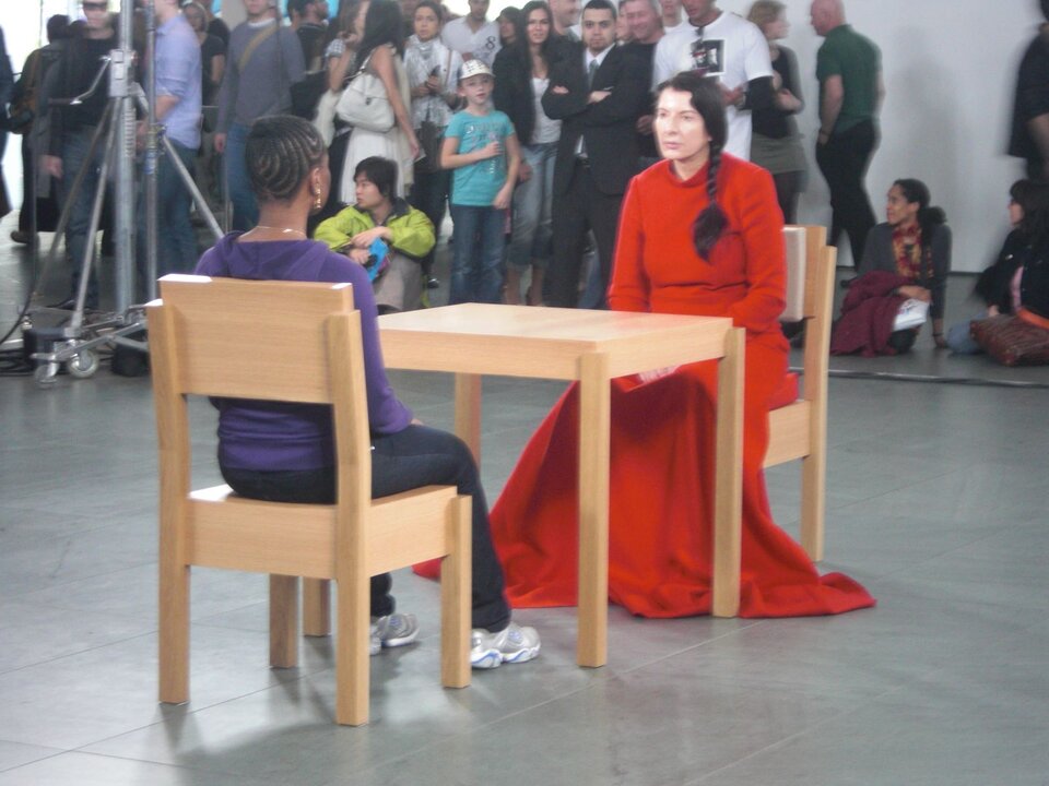 Ilustracja przedstawia performance Mariny Abramović "The Artist Is Present”. W niewielkim wnętrzu przy stole, naprzeciw siebie siedzą na krzesłach: przodem do widza artystka i tyłem - kobieta. Obie postaci są jakby zastygłe w bezruchu i wpatrują się w siebie. W tle widoczna jest grupa ludzi przyglądających się siedzącym.