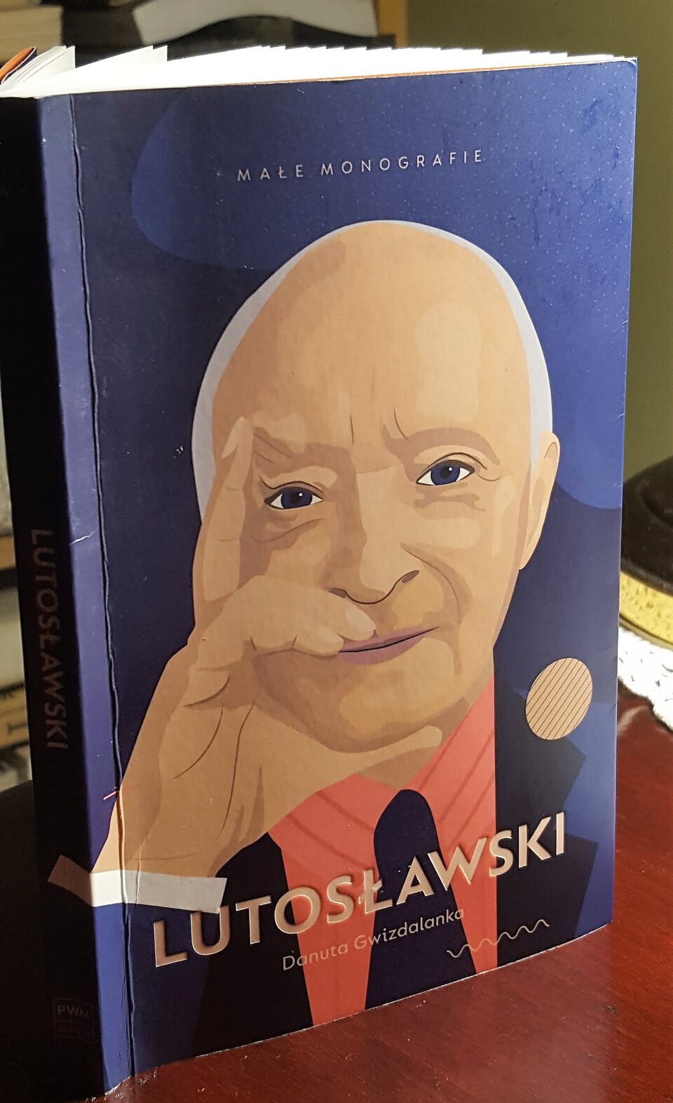 Na zdjęciu znajduje się stojąca na stole książka. Na okładce książki znajduje się starszy mężczyzna ubrany w niebieską marynarkę, czerwoną koszulę oraz czarny krawat. Mężczyzna prawą ręką dotyka swojej twarzy. Książka jest w kolorze granatowym. W dolnej części okładki znajduje się tytuł oraz autor.