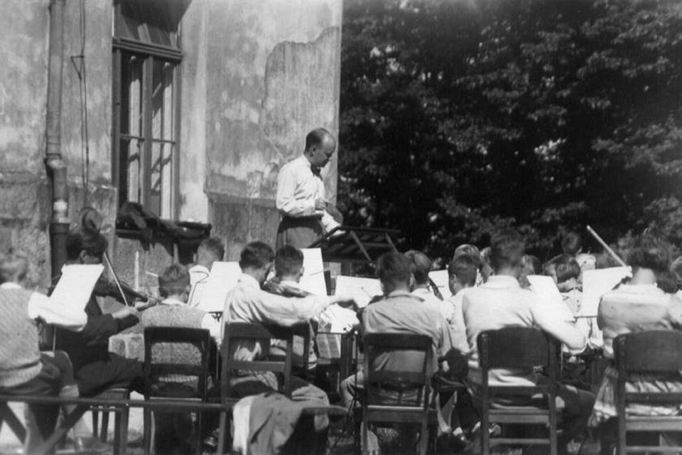 Ilustracja przedstawia Hindemith'a podczas próby koncertu w Plön. Mężczyzna na świeżym powietrzu szkoli młodych chłopców, którzy tyłem są odwróceni do fotografa. Chłopcy ubrani są w białe koszule, grają na instrumentach.