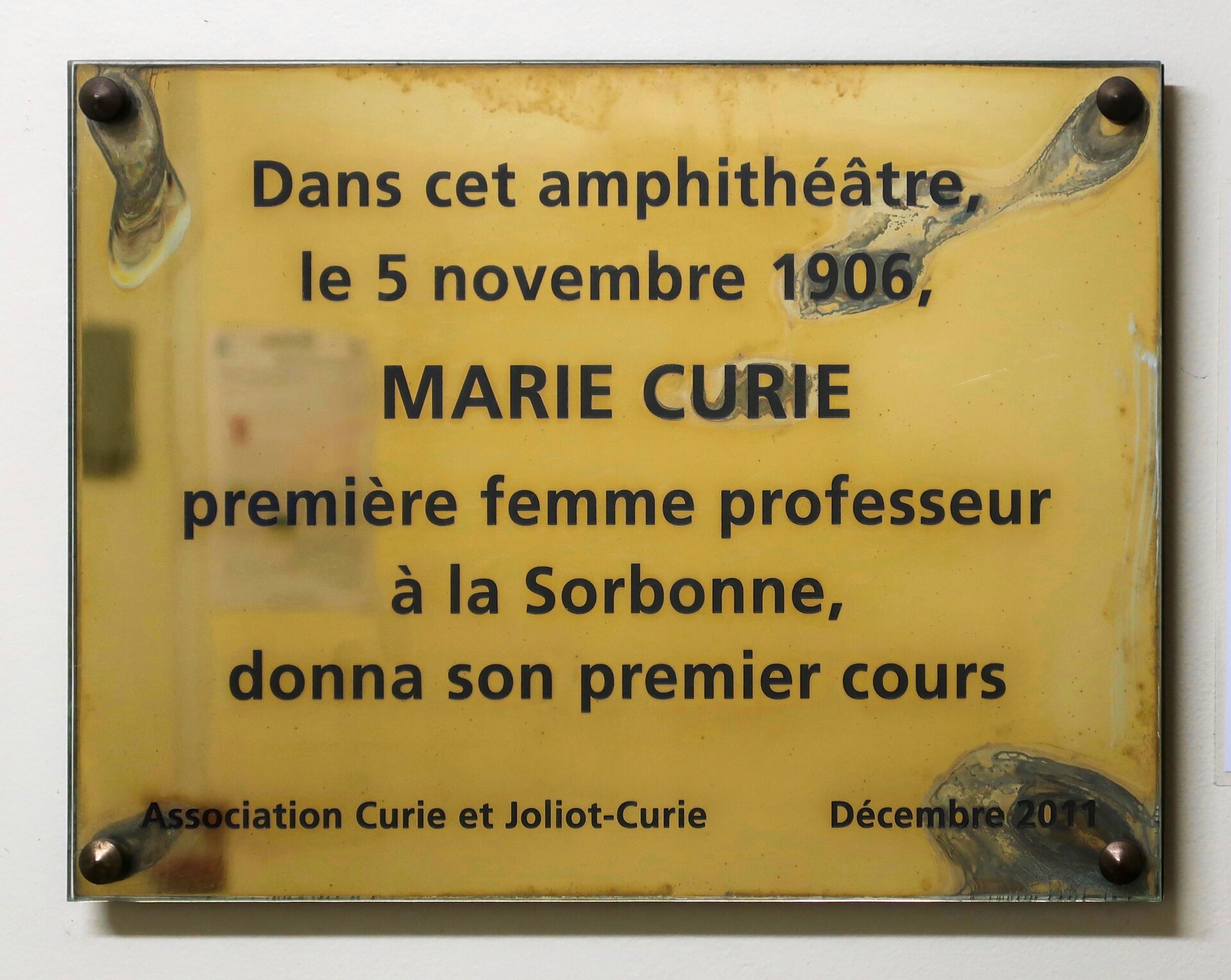 Na zdjęciu widać złotą tabliczkę z czterema śrubami, w rogach, które przytwierdzają ją do ściany na tabliczce umieszczono napis:  Dans cet amphithéâtre, le 5 novembre 1906, Marie Curie première femme professeur à la Sorbonne, donna son premiers cours. Association Curie et Joliot‑Curie. Decembre 2011. 