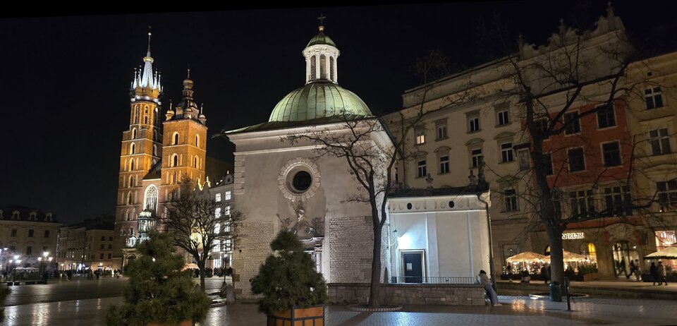 Fotografia przedstawia fragment Rynku Głównego w Krakowie. Na pierwszym planie kościół św. Wojciecha. Z lewej strony Sukiennice, a w oddali Kościół Mariacki