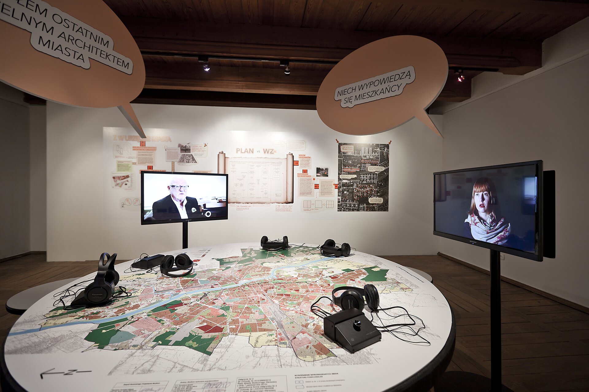 Na zdjęciu znajduje się pomieszczenie o brązowym suficie, brązowej podłodze i białych ścianach. Na środku pomieszczenia stoi okrągły stół. Na blacie przedstawiony jest plan miasta i leży na nim 5 czarnych zestawów z słuchawkami. Po prawej stronie na długiej, czarnej nodze stoi ekran. Wyświetlone jest na nim popiersie kobiety o rudych włosach. Po lewej stronie stoi drugi ekran. Wyświetlone jest na nim popiersie starszego mężczyzny w garniturze i okularach. Nad każdym ekranem wisi beżowy dymek z czarnym napisem. W tle są plakaty i plany powieszone na ścianie. Na suficie jest czarna szyna i z małymi lampkami. 