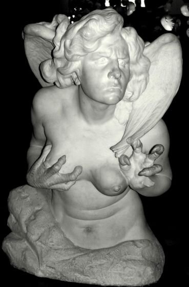 Czarnobiała fotografia przedstawia rzeźbę wykonaną z jasnego kamienia. Przedstawia popiersie półnagiej postaci, która ma głowę i ciało ludzkie oraz skrzydła i pazury ptaka. Ma bujne kręcone włosy. Jej lewa dłoń skierowana jest do przodu, a prawą dłonią trzyma swoją pierś. Rzeźba ustawiona jest na czarnym tle.