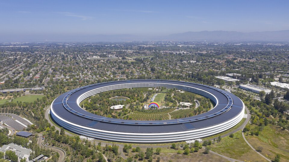 Zdjęcie przedstawia Apple Park, główną siedzibę firmy Apple Inc. w Dolinie Krzemowej. Budowla ma kształt wielkiego okręgu. We wnętrzu okręgu znajdują się tereny zielone, staw, park z kolorową tęczą pośrodku.