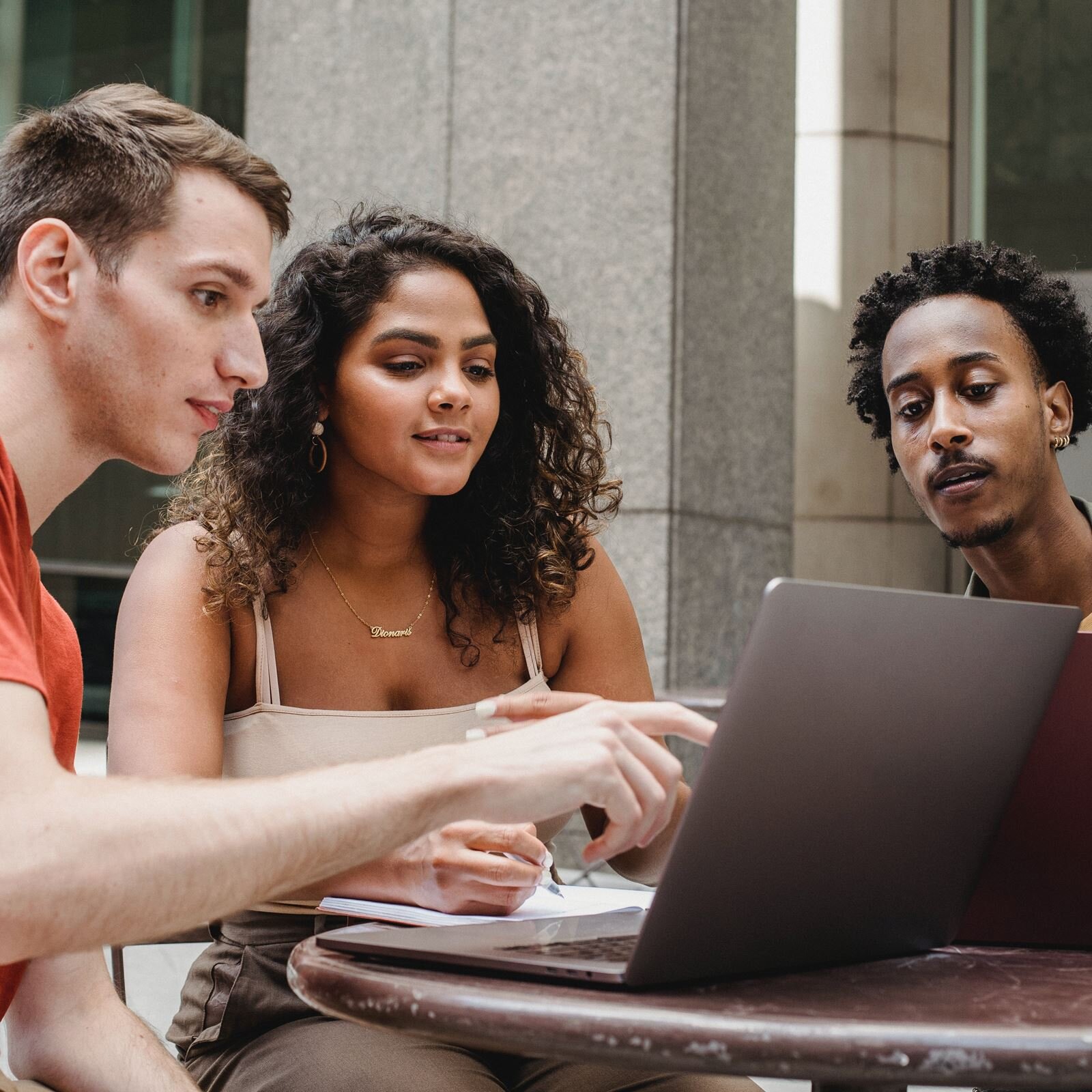 Zdjęcie przedstawia troje młodych ludzi różnych narodowości siedzących przy stoliku, na którym stoi otwarty laptop. Wszyscy się w niego wpatrują. 