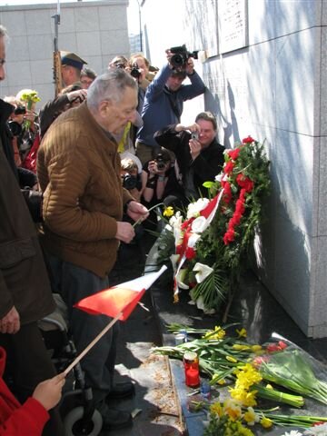 Zdjęcie przedstawia mężczyznę w średnim wieku, widzianego z profilu. Ma krótko ostrzyżone, gładkie włosy. Są zaczesane do góry. Mężczyzna ma pociągłą twarz i wyraźnie zarysowane brwi. Ma także duży nos i wydatne usta. Mężczyzna ubrany jest w kraciastą koszulę. A na nią założony ma sweter. Kładzie żółty żonkil pod pomnik, pod ścianę, pod którą leżą kwiaty i wieńce. Za nim są fotoreporterzy.
