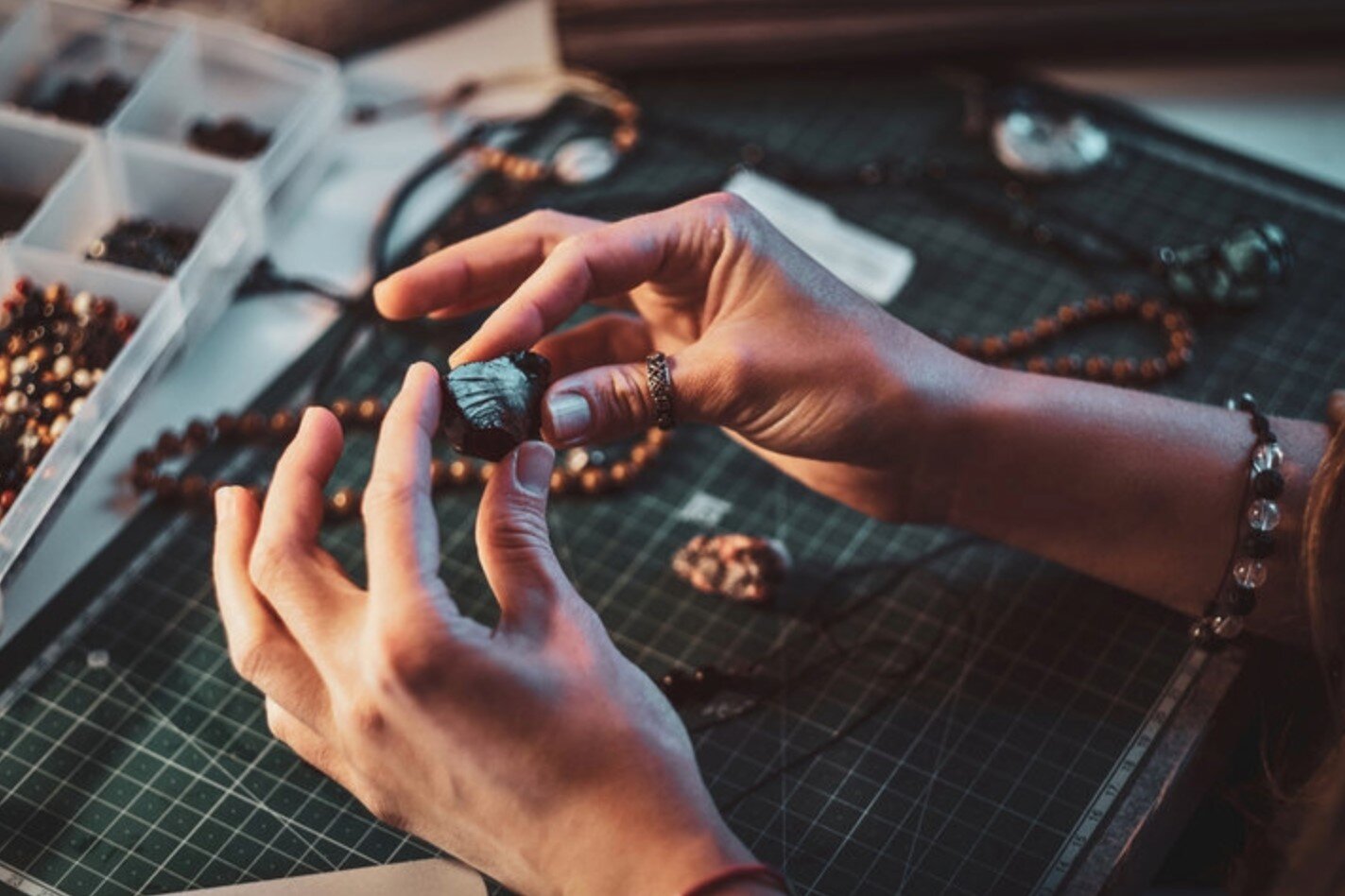 Ilustracja przedstawia fotografię dłoni opartych o blat stołu, na którym leżą rzemyki i koraliki ułożone w pojemnikach - znajdujących się w kadrze tylko częściowo. Ręce są sfotografowane podczas pracy:  w palcach znajduje się muszelka, która prawdopodobnie zostanie wykorzystana do stworzenia ozdoby.