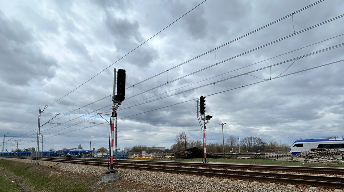 Ilustracja przedstawia dwa semafory wjazdowe. Semafory wjazdowe znajdują się obok torowiska. Semafory wjazdowe są to biało czerwone słupy, na których znajdują się lampy z pięcioma światłami, światła są wyłączone. Światła umiejscowione są w sposób pionowy, jedno pod drugim. Nad torowiskiem znajdują się linie napięcia a w tle przejeżdżający pociąg koloru biało niebieskiego.