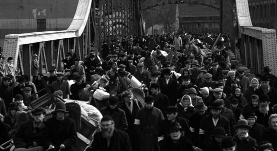 Zdjęcie przedstawia most, przez który przechodzi tłum ludności żydowskiej z opaskami na prawych ramionach. Ludzie niosą tobołki, torby, walizki.