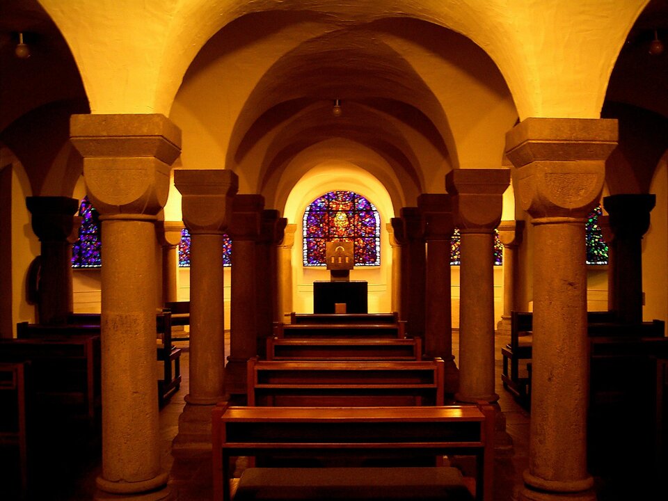 Fotografia przedstawia wnętrze kolegiaty św. Patroclusa w Soest w Niemczech. Na pierwszym planie znajduje się nawa, wzdłuż której biegną rzędy masywnych kolumn. Trzon kolumny jest w kształcie walca, natomiast głowica kształtem przypomina kielich, który rozszerza się w górę aż do prostopadłościennego zwieńczenia. Nad kolumnami widoczne są łuki łączące się na stropie w sklepienie. Pomiędzy kolumnami znajdują się drewniane ławki dla wiernych, a na końcu rzędu znajduje się prosty ołtarz i tabernakulum, nad którym widoczny jest kolorowy witraż. 