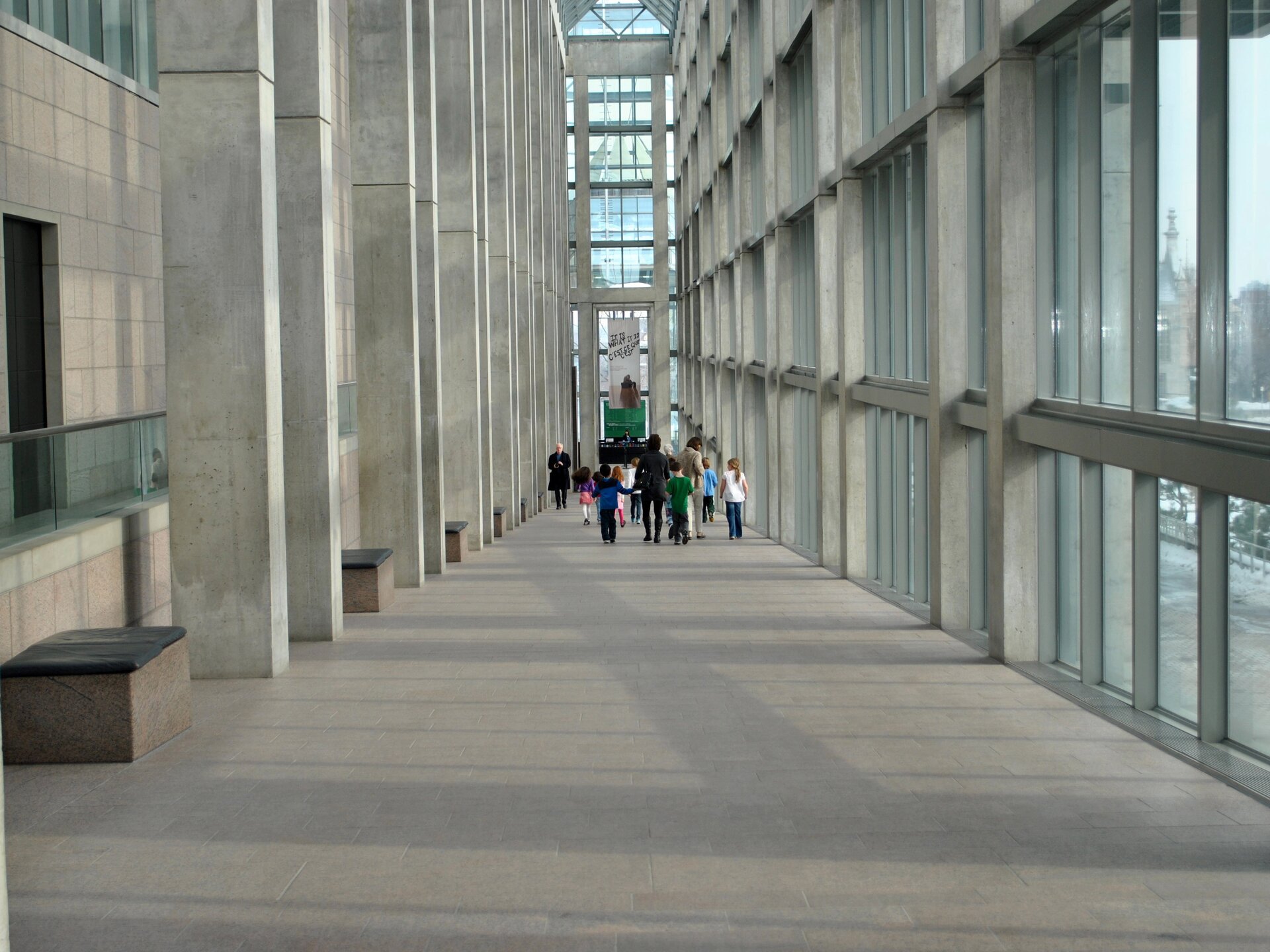 Fotografia barwna przedstawiająca wewnętrzny korytarz Narodowej Galerii Kanady w Ottawie. Po lewej filary z jasnego granitu podtrzymujące wysoką konstrukcję budynku, między nimi wyściełane ławki. Po prawej i naprzeciw nas przeszklone ściany. W głębi korytarza, odwrócone tyłem, dwie kobiety z grupką dzieci. Przed nimi idący ku nam mężczyzna w czarnym ubraniu. 