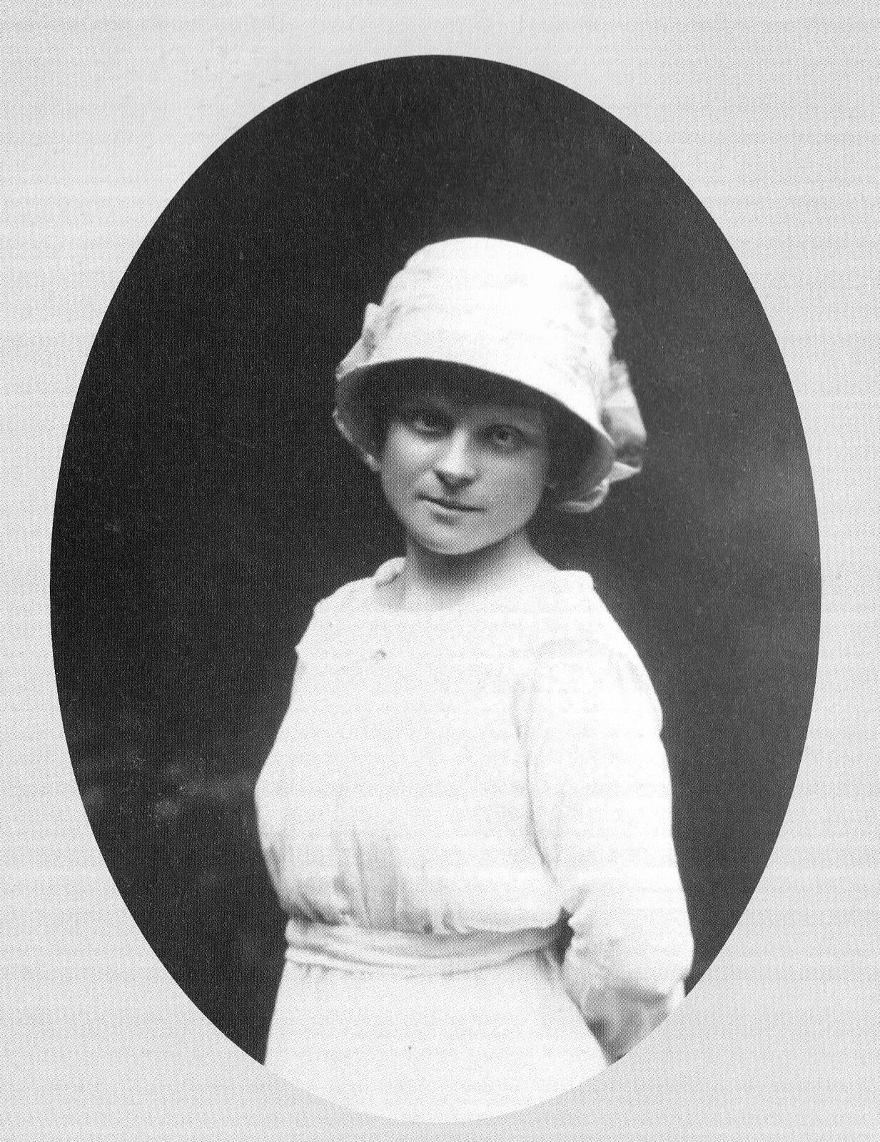 Czarno‑białe zdjęcie przedstawia portret młodej kobiety o delikatnej twarzy i ciemnych włosach ukrytych pod białym, materiałowym kapeluszem. Kobieta nosi białą sukienkę z długimi rękawami, zebraną w pasie białym paskiem. Tło jest ciemne.