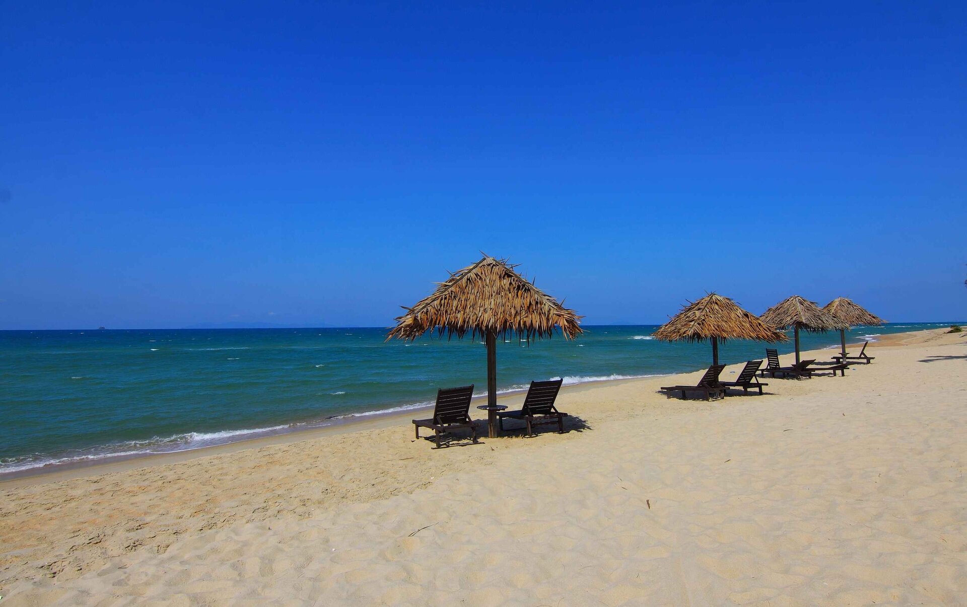 Rys. 2. Zdjęcie przedstawia fragment nadmorskiej plaży z leżakami, nad którymi ustawiono drewniane parasole pokryte liśćmi. Zdjęcie można podzielić na trzy poziome plany kolorystyczne: błękit nieba, turkusowe wody morza i bardzo jasny w kolorze piasek.