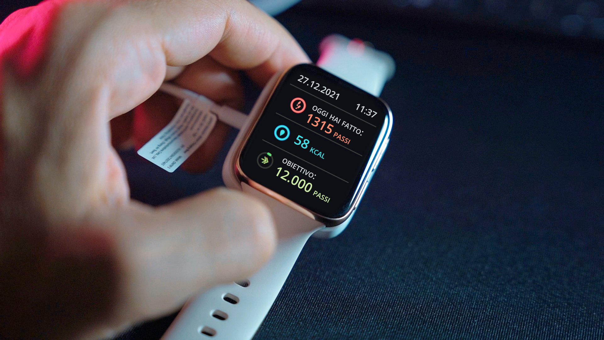 Zdjęcie przedstawia zbliżenie na zegarek typu smartwatch. Na ekranie zegarka znajduje się raport dzienny. Na górze ekranu zegarka, po lewej stronie widoczna jest data: 27.12.2021, a po prawej godzina: 11:37. Poniżej w pierwszej linijce znajduje się piktogram przedstawiający odciśnięte stopy oraz napis: Oggi hai fatto: 1315 passi. W drugiej linijce znajduje się piktogram przedstawiający kroplę oraz napis: 58 kcal. W trzeciej linijce widoczny jest piktogram przedstawiający strzałkę oraz napis: Obiettivo: 12000 passi.