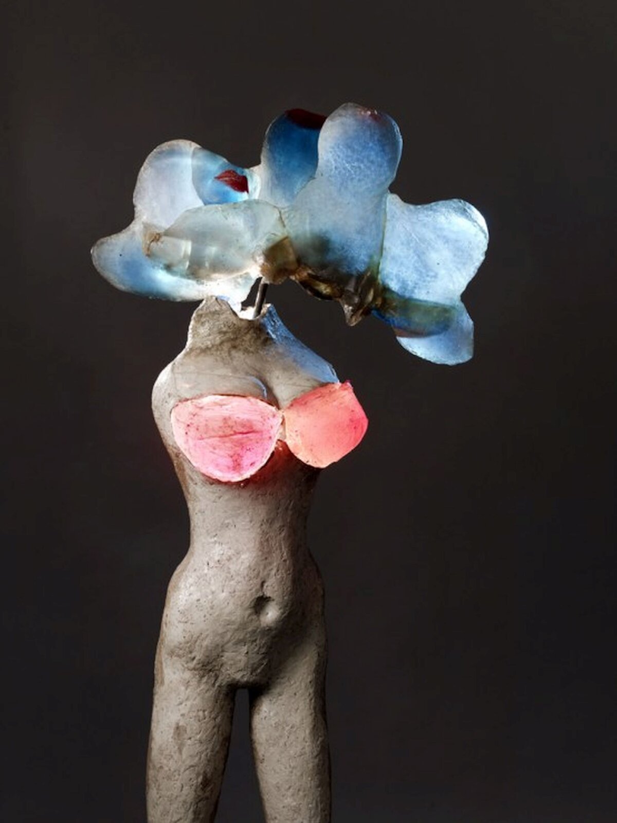 Zdjęcie przedstawia rzeźbę Aliny Szapocznikow Iluminowana. Naga postać kobiety, bez rąk, piersi ma schowane w czerwonym biustonoszu. Zamiast głowy świecący na niebiesko element o kształcie przypominającym meduzę.