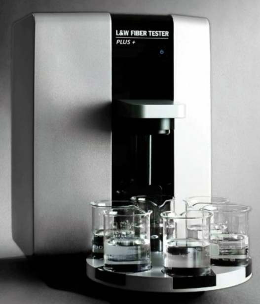 Na zdjęciu znajduje się tester włókien, który wyglądem przypomina ekspres do kawy. Na jego środku znajduje się wypustka stanowiąca dozownik. Pod nią, na okrągłej podstawce, ułożono sześć szklanych zlewek.