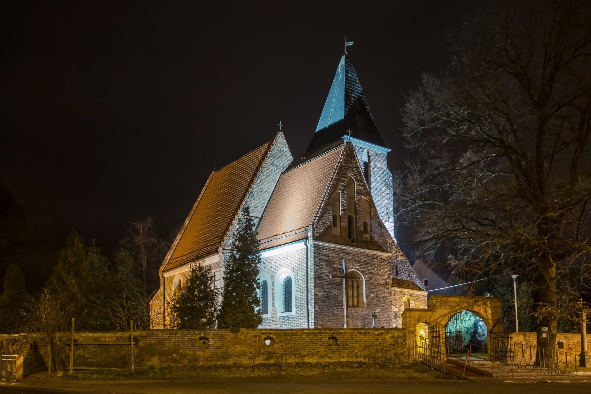 Zdjęcie współczesne przedstawia widok na kościół parafialny, oświetlony sztucznym światłem o zmierzchu. Na pierwszym planie widoczne zewnętrzne ściany prezbiterium, w głębi nawa o wyższym dachu, oba elementy konstrukcyjne znajdują się pod dwuspadowym dachem. W głębi, bliżej prawej krawędzi zdjęcia, widoczna dzwonnica, z zadaszeniem w kształcie ostrosłupa. Kościół jest otoczony murem, prezentowanym na zdjęciu przez całą jego szerokość. Między świątynią a murem znajduje się drewniany krzyż. Bliżej prawej krawędzi zdjęcia widoczna brama prowadząca na teren świątyni, przed nią drewniana kładka. Bezlistne drzewo po prawej stronie, drzewa iglaste w centrum zdjęcia, bliżej prezbiterium, w tle pozostałe drzewa, bliżej lewej krawędzi. 