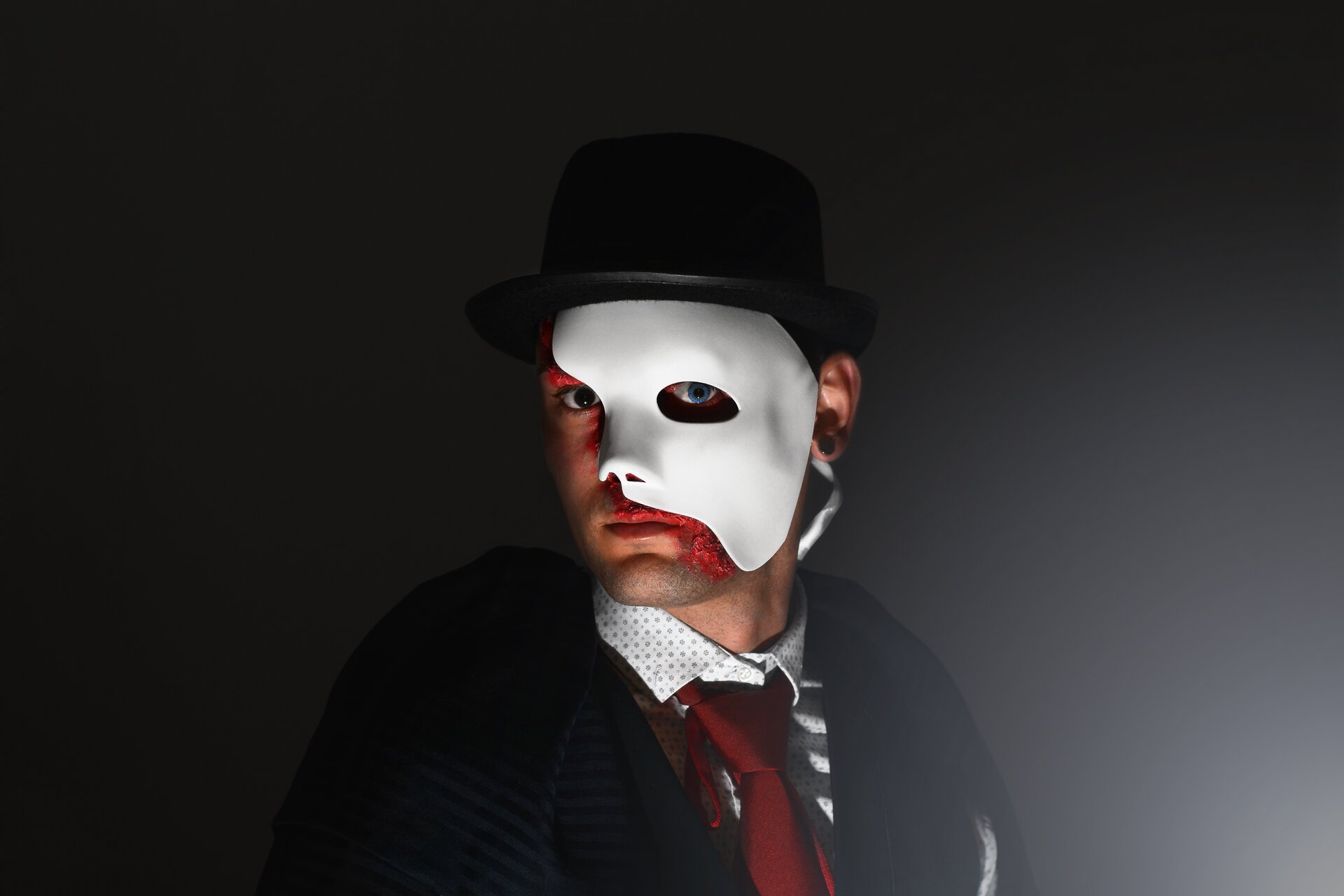 Ilustracja przedstawia zdjęcie aktora wcielającego się w postać musicalową. Na ciemnym tle widnieje portret mężczyzny w meloniku z białą maską, zasłaniającą połowę twarzy. Postać ubrana jest w ciemny płaszcz, jasną koszulę oraz czerwony krawat.