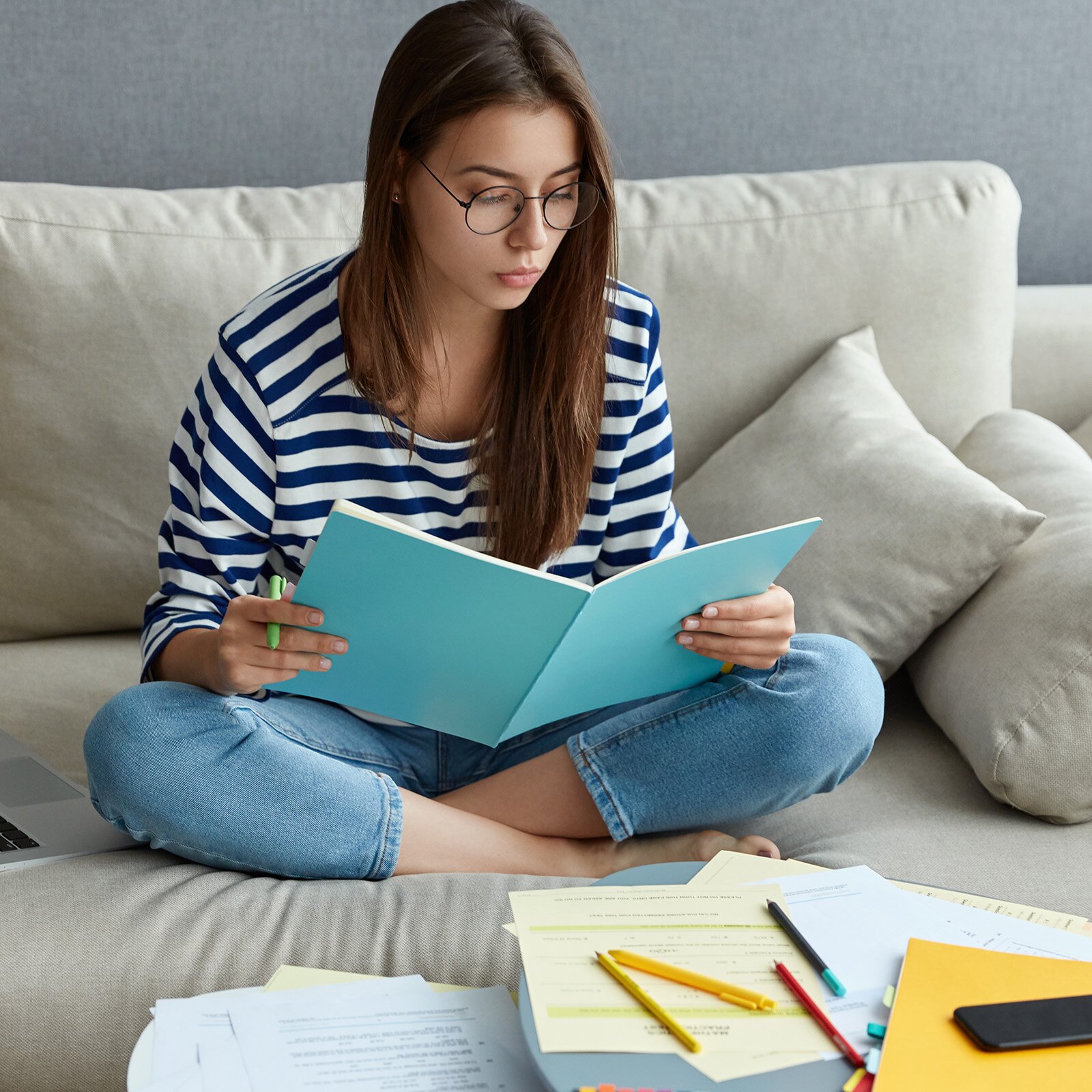 Zdjęcie przedstawia młodą dziewczynę w okularach ok lat szesnastu siedzącą na sofie. Dziewczyna trzyma w rękach zeszyt i długopis. Wzrok ma opuszczony w dół. Przed nią stoi mały stolik, na którym rozłożone są kartki z notatkami, kolorowe długopisy i flamastry, zeszyt ze znacznikami i telefon komórkowy.