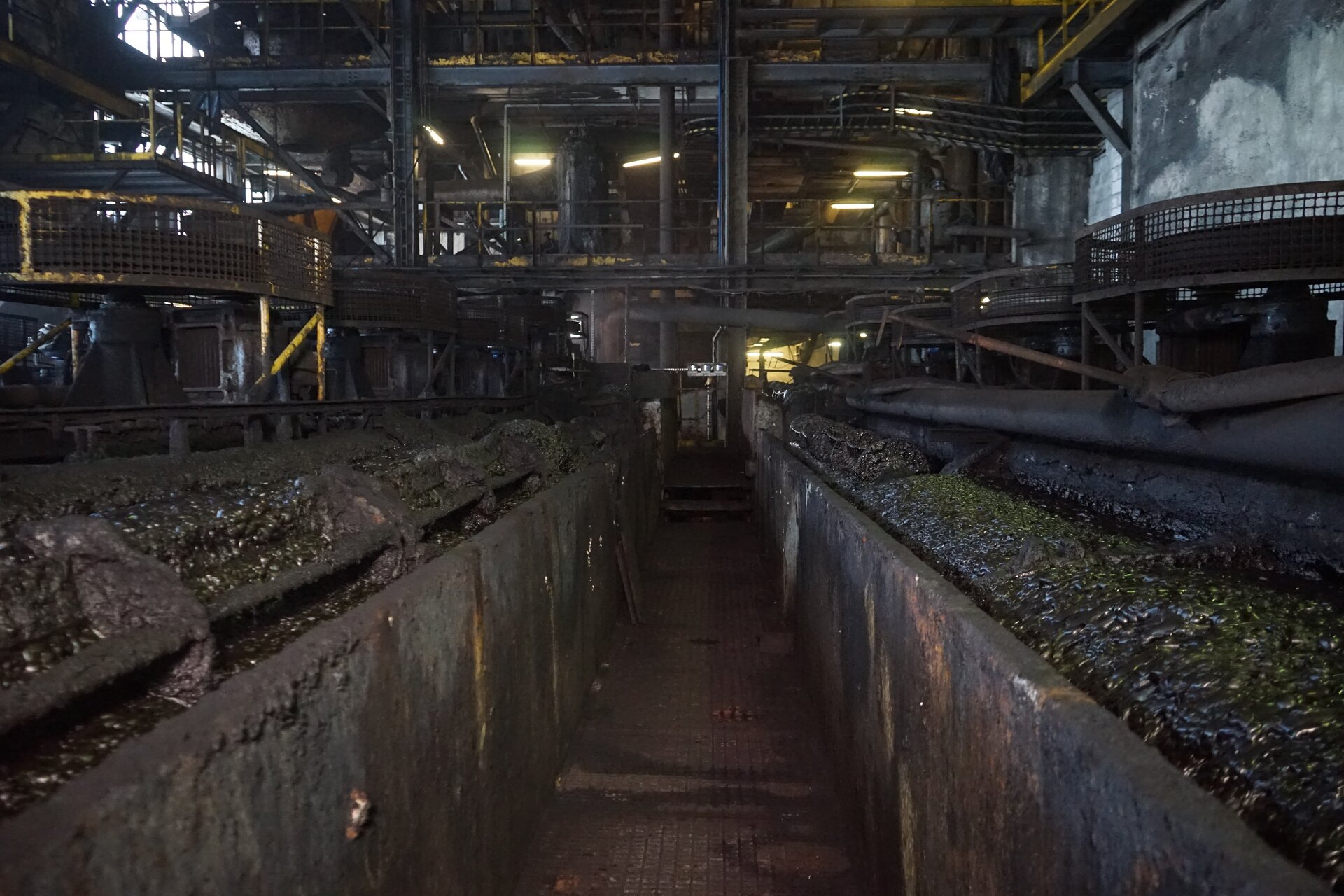 Na zdjęciu widoczna jest maszyna flotacyjna. Maszna flotacyjna jest to urządzenie umożliwiające wzbogacanie fizykochemiczne za pomocą metody flotacyjnej. Na zdjęciu widoczna jest hala przemysłowa. Po obu stronach zdjęcia znajdują się podłużne prostopadłe stalowe pojemniki. Pojemniki są wypełnione czarną cieczą. Ciecz jest zbliżona konsystencją do szlamu. Pomiędzy pojemnikami jest przejście. Nad pojemnikami znajdują się stożkowe kształty. Na ich szczycie osadzone są okrągłe obiekty. Po obwodzie obiektów biegnie siatka.