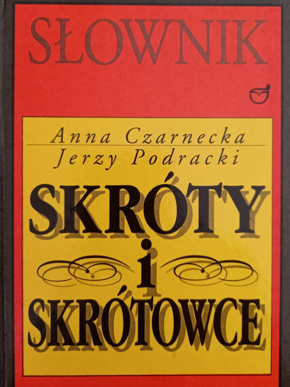 Kolorowe zdjęcie przedstawia okładkę "Słownika skrótów i skrótowców". W czerwonym prostokącie umieszczono mniejszy, żółty kwadrat. Na czerwonym tle napis czarnymi literami: Słownik. Poniżej na żółtym tle imiona i nazwiska autorów słownika: Anna Czarnecka, Jerzy Podracki. Poniżej pogrubionym drukiem duży napis czarnymi literami: SKRÓTY I SKRÓTOWCE.