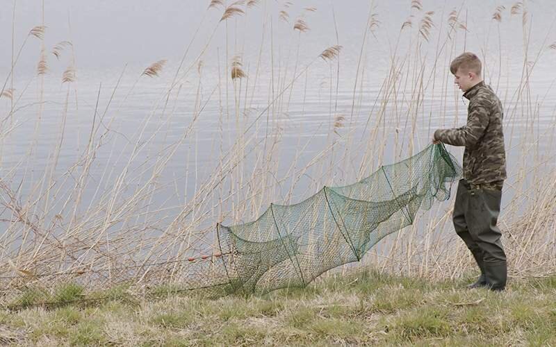 Krok pierwszy, podpisany Przygotowanie sieci. Na zdjęciu widać mężczyznę ubranego w spodnie koloru khaki i bluzę moro. Stoi na brzegu zbiornika wodnego i rozwija zieloną sieć rybacką.