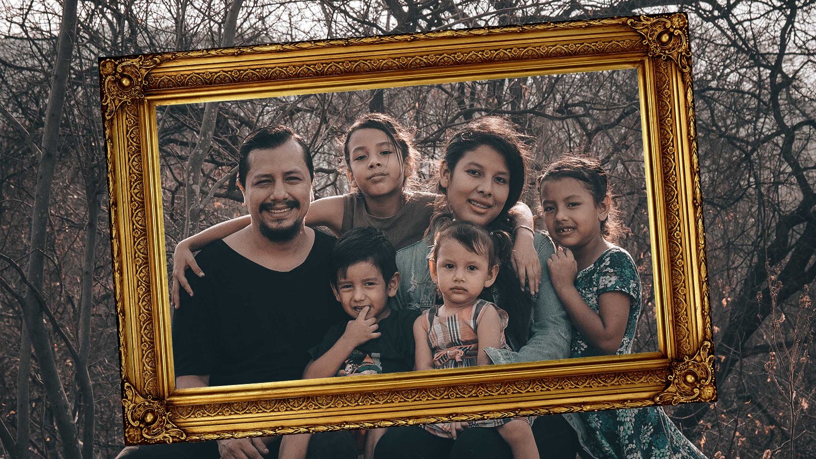 Zdjęcie przedstawia grupkę ludzi, dorosłego mężczyznę oraz kobietę i czworo dzieci, dwie dziewczynki w wieku szkolnym i dwoje przedszkolaków. Rodzinne zdjęcie jest w złotej ramie na tle drzew bez liści.