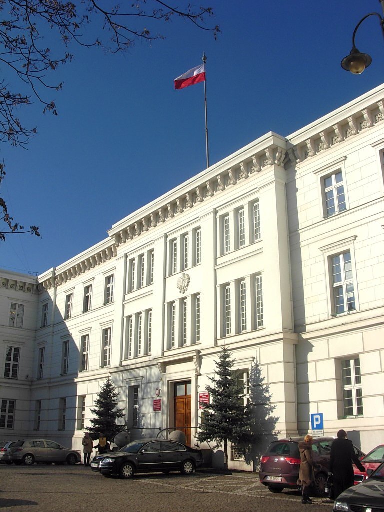 Kliknij aby powiększyć Zdjęcie przedstawia budynek w stylu klasycystycznym kolorze białym. Na dachu widoczny jest maszt, na którego szczycie powiewa flaga w barwach biało‑czerwonych. Parter budynku zdobiony jest boniowaniem, front główny zwieńczony jest gzymsem, nad którym znajduje się płaskorzeźba orła. Przed budynkiem rozciąga się parking, na którym stoją zaparkowane samochody. Przed wejściem do budynku ustawione zostały dwie choinki.