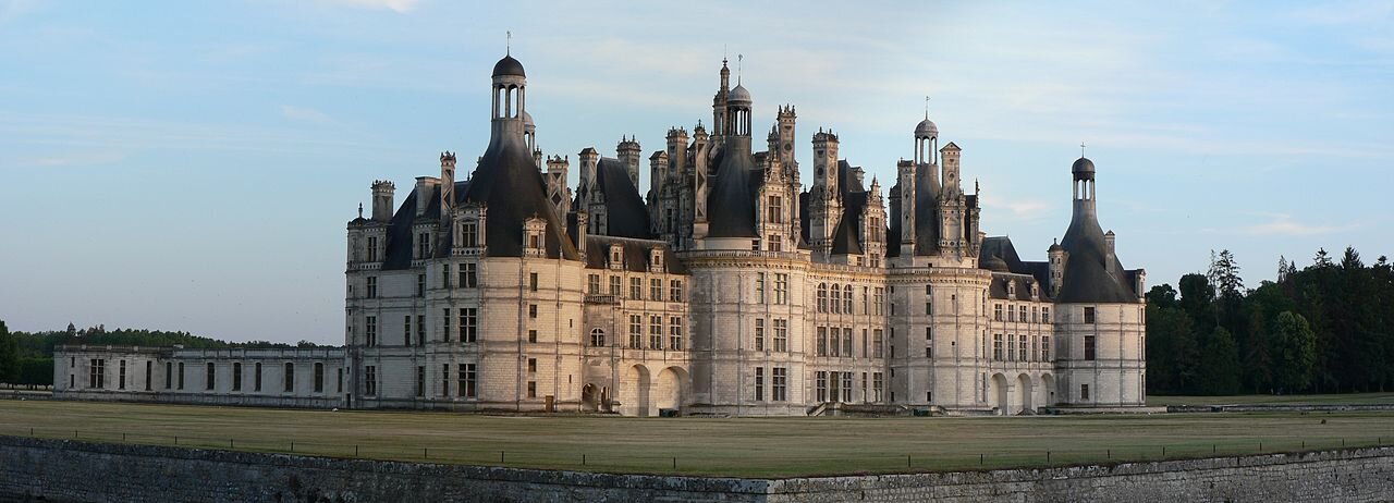 Widok renesansowego zamku w Chambord o planie nawiązującym do gotyckich zamków obronnych. Zamek jest w kształcie prostokąta, trzykondygnacyjny, otoczony fosą, posiadający na rogach wieże. Cztery wieże fasady zwieńczone są spadzistymi dachami z kampanilami (dzwonnicami). Fosa przed zamkiem ma wybetonowane brzegi. Niebo w tle jest pogodne.