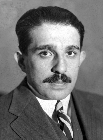 Czarno‑białe zdjęcie przedstawia popiersie mężczyzny w średnim wieku w garniturze. Ma czarne, krótkie włosy, odstające uszy, krzaczaste brwi i wąsy. Jego twarz jest poważna.

